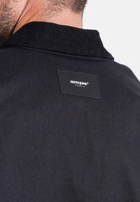 Giacca nera con colletto in velluto a coste e un tessuto strutturato. Presenta un'etichetta rettangolare nera con la scritta "SIXTH JUNE PARIS".