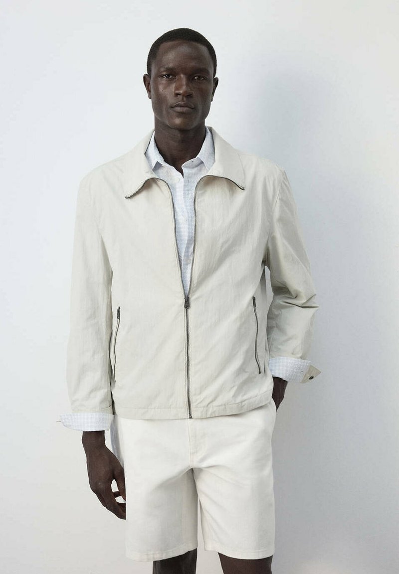Homme portant une veste beige clair zippée et un short assorti, debout devant un fond blanc uni.
