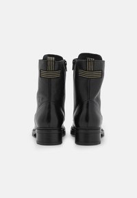 Botas de tobillo de cuero negro vistas desde atrás, con tiradores de crema cosidos en horizontal y vertical, y tacones bajos apilados, sobre un fondo blanco.