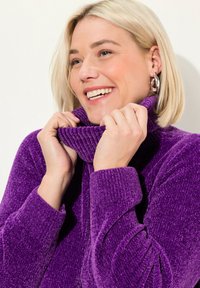 Maglione viola con collo alto realizzato in tessuto testurizzato, dotato di polsini a coste e un colletto accogliente, inquadra le mani che tengono il colletto.