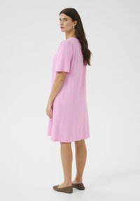 Kortärmad klänning i knälängd av ljusrosa tyg. Slät textur, något utsvängd design, med minimal dekoration. Skor: mjuka, bruna ballerinas.