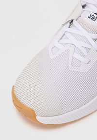 Close-up of white athletic shoe toe with textured mesh upper, white laces, and light brown rubber sole.