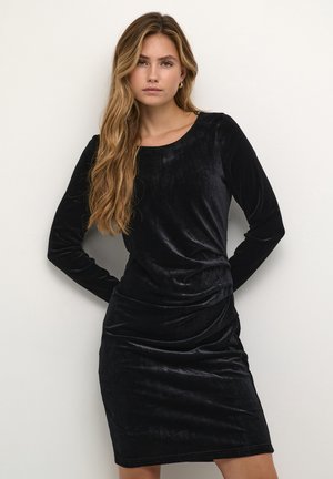 Robe longue en velours noir à manches longues avec un col rond, présentant une silhouette ajustée et une texture douce, accentuée par des détails froncés à la taille.