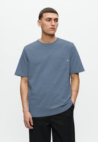 Camiseta de algodón azul con mangas cortas, cuello redondo y un bolsillo en el pecho. Presenta una textura sutil y un pequeño logotipo en el bolsillo.