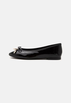 Ballerine noire brillante avec un petit nœud et un détail de lacet doré sur le bout arrondi, vue de profil sur un fond uni.