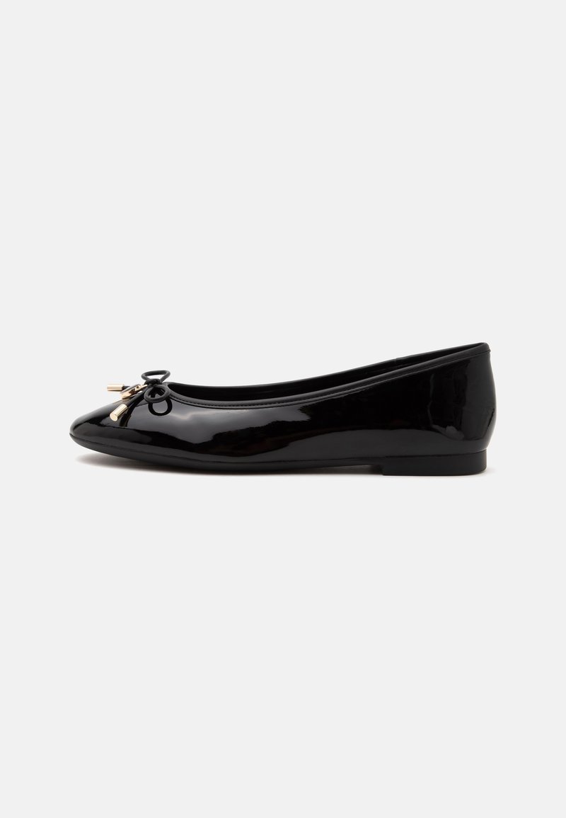 Ballerine noire brillante avec un petit nœud et un détail de lacet doré sur le bout arrondi, vue de profil sur un fond uni.