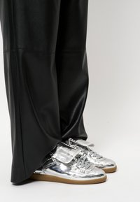 Metalen zilveren sneakers met een gladde textuur, voorzien van een klep over de veters, gecombineerd met losse zwarte leren broeken.