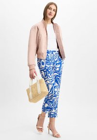 Vrouw in een roze bomberjack, witte top, blauw-witte gemusterde broek, naakthoge hakken en een geweven handtas.