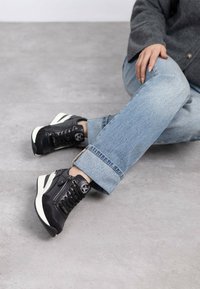 Baskets en cuir noir avec des accents texturés, semelles blanches et détail de fermeture éclair sur le côté. Associées à un jean bleu clair retroussé.