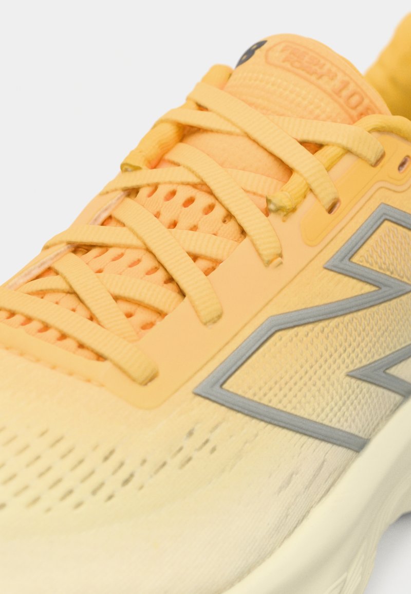 Zapatilla de correr amarilla y naranja con parte superior de malla, cordones entrecruzados y logo "N" gris en el lateral.