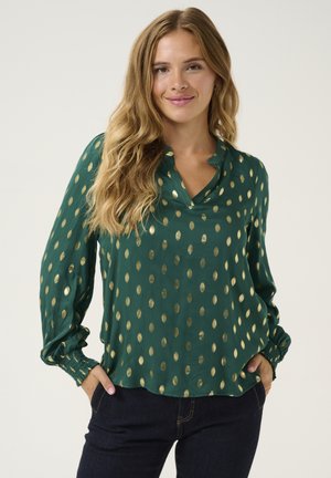 Green blouse with gold polka dots, soft fabric, v-neckline, long sleeves with gathered cuffs, relaxed fit, and slight sheen.