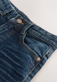 Közelkép kék farmernadrágról, amelyen jól látható az elülső zseb, az övhurkok és a réz szegecs részletei a texturált anyagon.