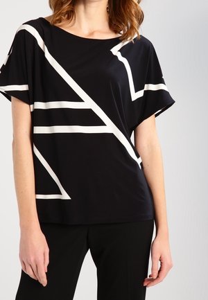 Vrouw draagt een zwart shirt met korte mouwen en witte geometrische lijnen, en een zwarte broek, zichtbaar van schouders tot heupen.