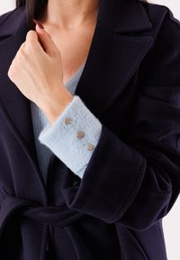 Donna che indossa un cappotto blu scuro sopra un maglione azzurro con polsino abbottonato, tiene la risvolto del cappotto vicino al collo con la mano sinistra.