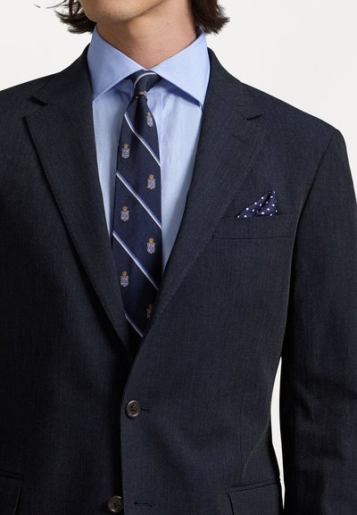 Veste de costume marine en tissu texturé, chemise habillée bleu clair, cravate rayée ornée d'emblèmes et pochette à pois.
