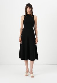 Svart ermeløs kjole med tettsittende overdel og utbrettelig skjørt, laget av et glatt stoff. Et par off-white sandaler med glid og trekantede detaljer.