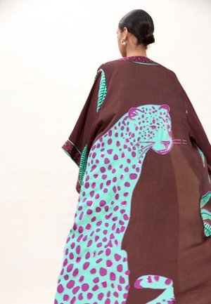 Mujer con cabello oscuro recogido en un moño, que lleva una capa marrón con un gran diseño de leopardo con manchas turquesas y moradas en la espalda.