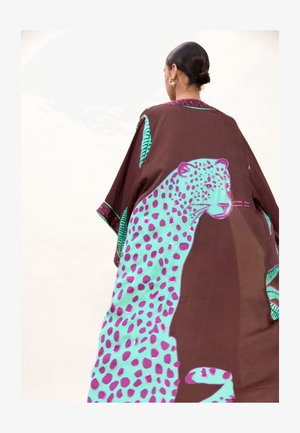 Mujer con cabello oscuro recogido en un moño, que lleva una capa marrón con un gran diseño de leopardo con manchas turquesas y moradas en la espalda.