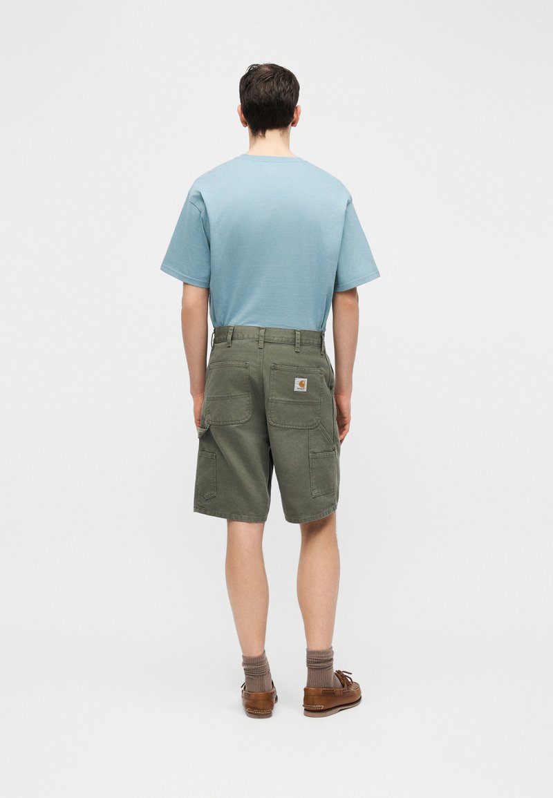 Homme debout de dos portant une chemise à manches courtes bleu clair, un short cargo vert olive avec des poches arrière, des chaussures bateau marron et des chaussettes grises.