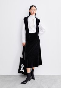 Schwarzer samtiger Midi-Rock kombiniert mit einem weißen Hemd mit Knöpfen und einer schwarzen Weste. Das Modell hält eine schwarze Tragetasche mit einem weißen Grafikdesign. Schwarze Absatz-Stiefeletten.