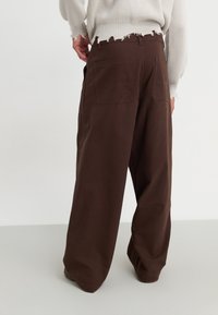 Pantalons brown à jambes larges en coton doux, avec deux poches arrière et un design d'ourlet brut et effiloché.
