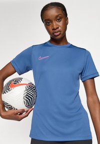 Μπλε αθλητική μπλούζα Nike με κοντά μανίκια και ροζ λεπτομέρεια λογότυπου, κρατημένη δίπλα σε μια ποδοσφαιρική μπάλα με μαύρο και άσπρο σχέδιο που διαθέτει πορτοκαλί λεπτομέρεια.