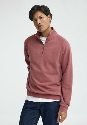 Uomo con capelli scuri che indossa una felpa rosa con mezza zip sopra una camicia bianca e jeans scuri, su uno sfondo chiaro e semplice.