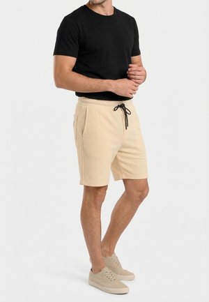 Hombre con camiseta negra de manga corta, pantalones cortos beige con cordón y zapatillas deportivas beige, de pie con las manos juntas al frente.
