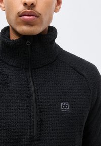 Close-up of a person wearing a black textured fleece jacket with a high collar and a front zipper, featuring a small "66° NORTH" logo.