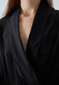 Blusa nera in un tessuto liscio, con un profondo scollo a V e un design frontale drappeggiato. Texture morbida con linee minimaliste e pulite.