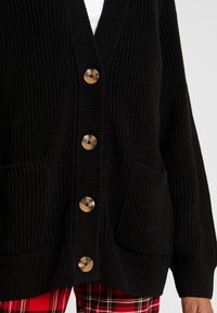 Černý pletený cardigan s výstřihem do V, šesti velkými kulatými knoflíky, dvěma předními kapsami a žebrovanými manžetami.