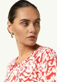 Blusa a fiori rossi e bianchi con scollatura a barco e dettagli con bottoni, realizzata in un tessuto morbido con una superficie liscia. Orecchini a cerchio dorati visibili.