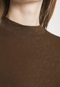 Bruine hooggesloten top van zachte, gestructureerde stof. Heeft een aansluitend ontwerp met geribde accenten rond de halslijn.