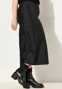 Schwarze, weite Cargo-Hosen mit glatter Textur, ausgestattet mit Seitentaschen und einem Reißverschlussdetail am Saum, ergänzt durch schwarze klobige Stiefeletten.