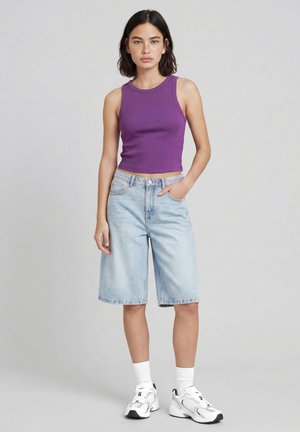 Jeune femme debout portant un haut sans manches violet, un short en jean bleu clair longueur genoux, des chaussettes blanches et des baskets blanches.