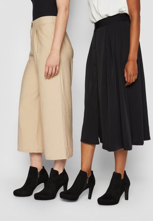 Beige plissierte Culottes und ein schwarzer Midi-Rock kombiniert mit schwarzen Wildleder-Ankle-Boots mit hohem Absatz und seitlichem Reißverschluss.
