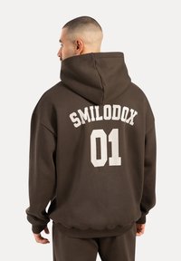Brauner übergroßer Hoodie mit einem großen weißen Druck auf dem Rücken, der "SMILODOX 01" liest. Verfügt über eine geräumige Kapuze und gerippte Bündchen.