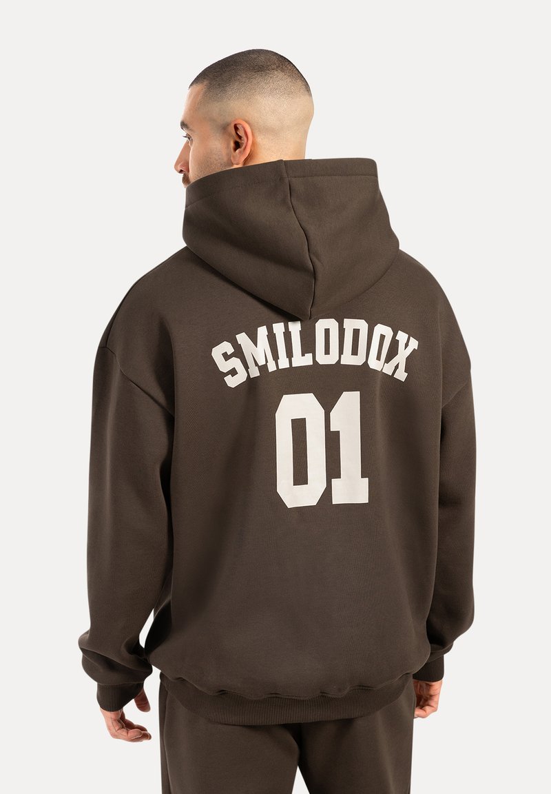 Brauner übergroßer Hoodie mit einem großen weißen Druck auf dem Rücken, der "SMILODOX 01" liest. Verfügt über eine geräumige Kapuze und gerippte Bündchen.