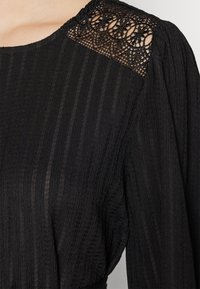 Μαύρο φόρεμα με ριμπ υφή και περίπλοκες δαντελένιες λεπτομέρειες στους ώμους. Μαλακό ύφασμα με χαλαρή εφαρμογή και μακριά μανίκια.