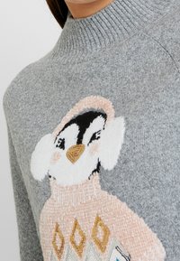 Grå stickad tröja med hög krage som har en pingvindesign i vitt, svart och rosa. Innehåller detaljerade geometriska mönster i guld och kräm.