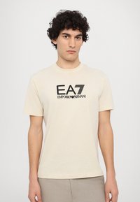 Béžové bavlněné tričko s černým logem EA7 Emporio Armani na přední straně. Krátké rukávy a kulatý výstřih. Měkká textura.