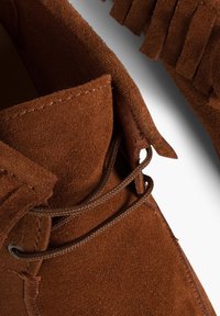 Bruine suède moccasinschoen met een franje kraag, ronde neus en bruine veters. De textuur is zacht met een matte afwerking.