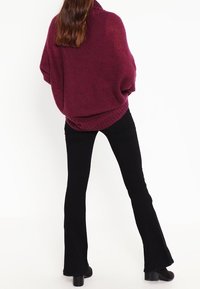 Dunkelroter, übergroßer Strickpullover mit hohem Kragen, langen Ärmeln und geripptem Saum, kombiniert mit schwarzen, ausgestellten Hosen und Stiefeln mit Absatz.