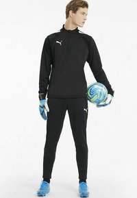 Černá atletická souprava s límcem na zip a bílým logem. Osoba drží modro-zelený fotbalový míč a má na nohou modré kopačky.