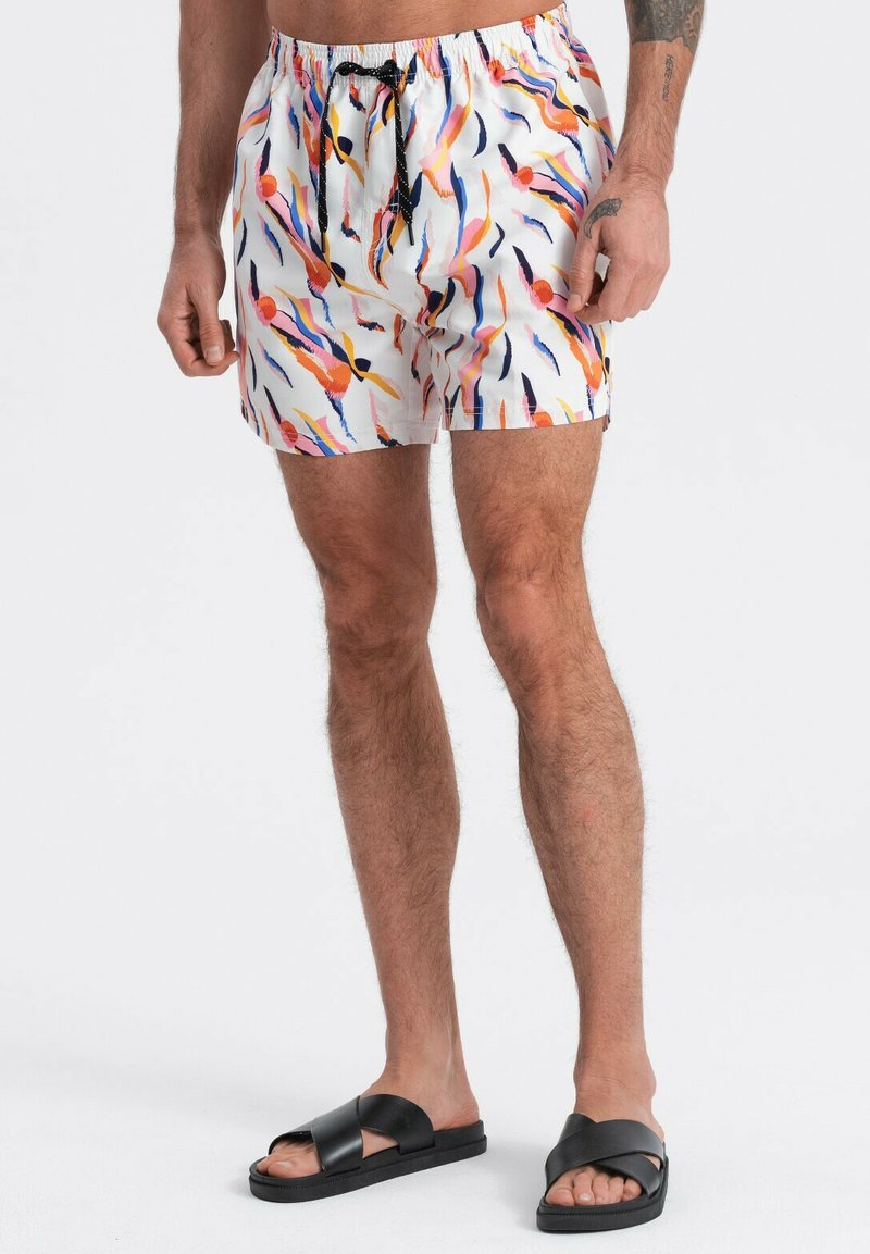 Homme portant un short de bain blanc avec des coups de pinceau abstraits colorés et des sandales noires à glissière, debout devant un fond blanc.