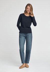 Granatowa koszula z długim rękawem, okrągłym dekoltem i małym czerwonym logo, zestawiona z jasnoniebieskimi jeansami o prostych nogawkach oraz brązowymi płaskimi butami.