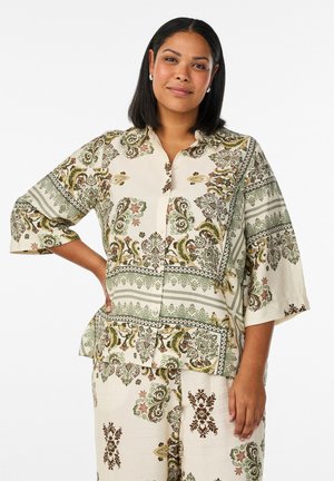 Naine seisab, üks käsi puusal, kannab sobivat kreemivärvi riietust, millel on rohelised ja pruunid keerukad lille- ja paisleimustrid.