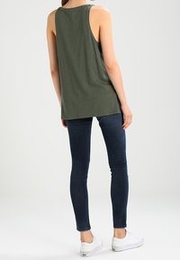 Olivgrön ärmlös topp, avslappnad passform, tillsammans med mörkblå skinny jeans och vita sneakers, sedd bakifrån. Enkelt design.