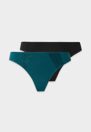Femme debout pieds nus en soutien-gorge noir sans bretelles et sous-vêtements bikini sur fond uni clair.