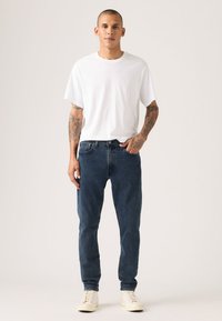 Balti trumpomis rankovėmis marškinėliai, mėlynos spalvos džinsai iš džinso audinio ir smėlio spalvos sportbačiai. Džinsai turi klasikinį kirpimą, džinso tekstūrą ir subtilų siuvimą.